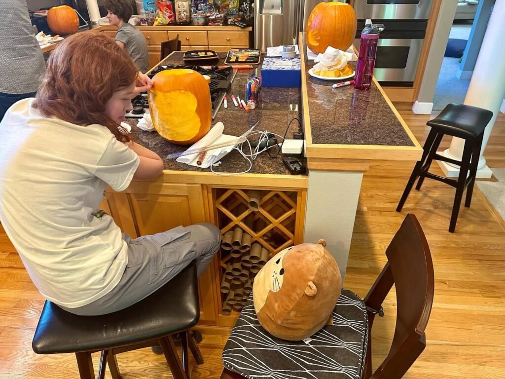 A child sits at a kitchen island carving a large orange pumpkin next to a stuffed toy.