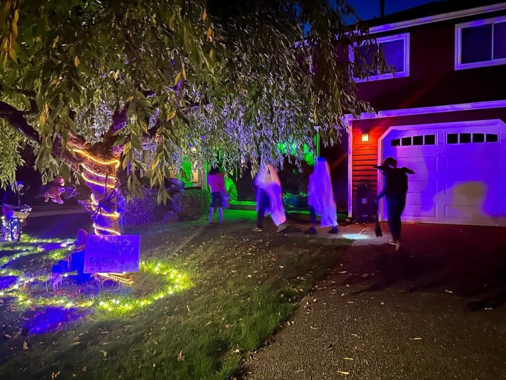 A two-story house illuminated by vibrant purple and blue Christmas lights, featuring a tree in the yard.