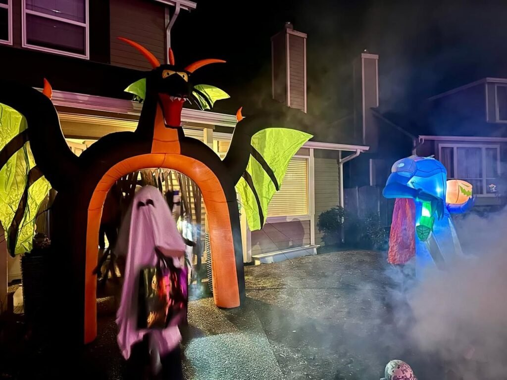 A close-up of a person in a ghost costume standing near an inflatable demonic arch. Halloween decorations and fog effects are visible in the nighttime setting.