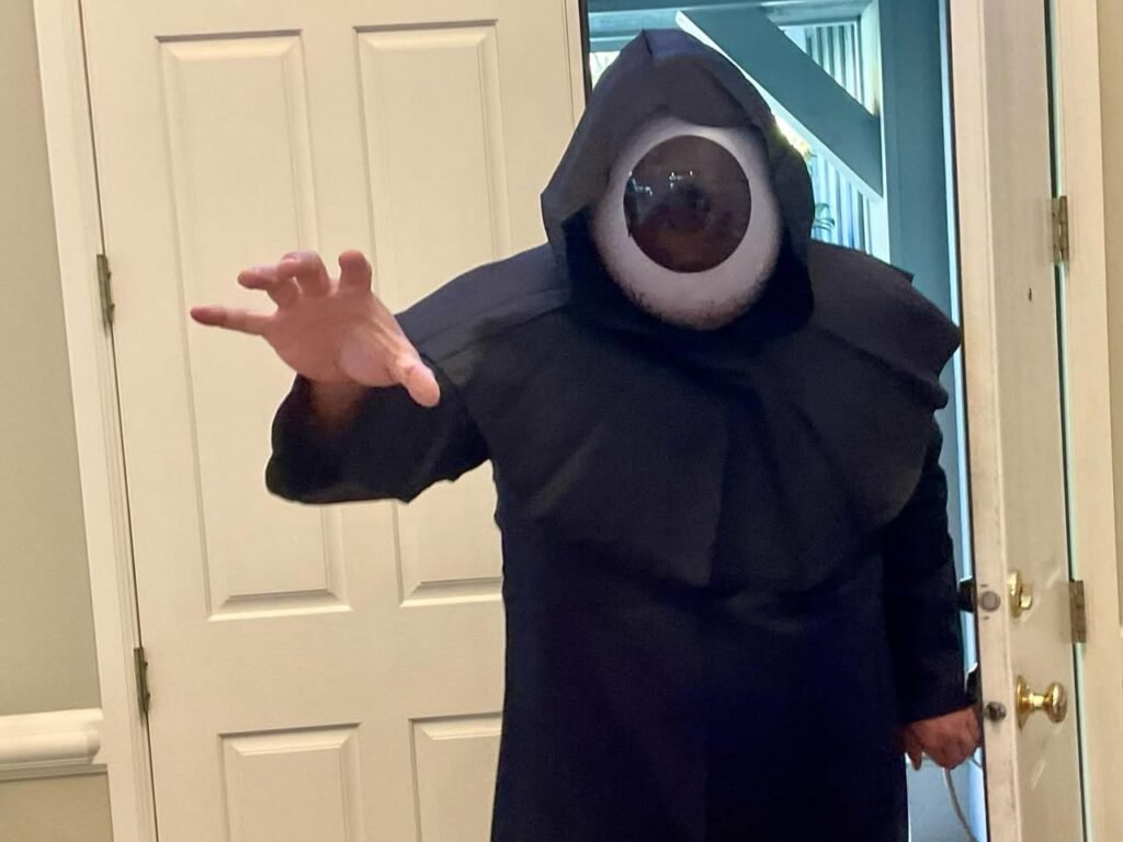 Person wearing a black hooded costume with a large eye mask, standing in a doorway with one arm extended.