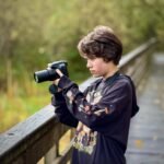 A teen takes photos on a wooden walkway. Foliage creates a soft, natural background.