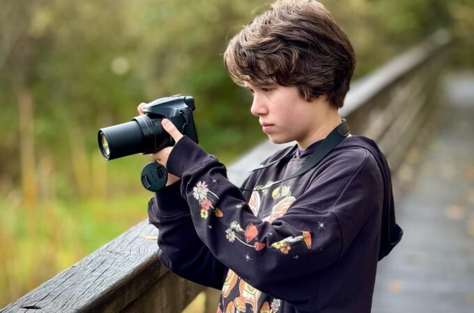A teen takes photos on a wooden walkway. Foliage creates a soft, natural background.