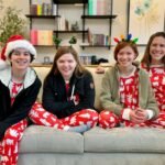 A cozy family poses in matching red and white Christmas pajamas on the couch. One person wears a Santa hat while others have festive headbands with reindeer antlers and lights.