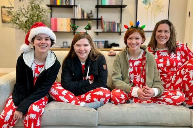 A cozy family poses in matching red and white Christmas pajamas on the couch. One person wears a Santa hat while others have festive headbands with reindeer antlers and lights.