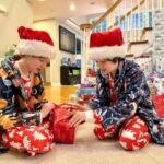 Macklemore Two people wearing Santa hats and festive pajamas opening a gift in a home. Gift wrapping paper is stacked nearby.