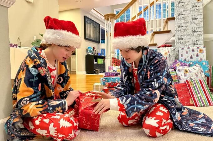 Two people wearing Santa hats and festive pajamas opening a gift in a home. Gift wrapping paper is stacked nearby.