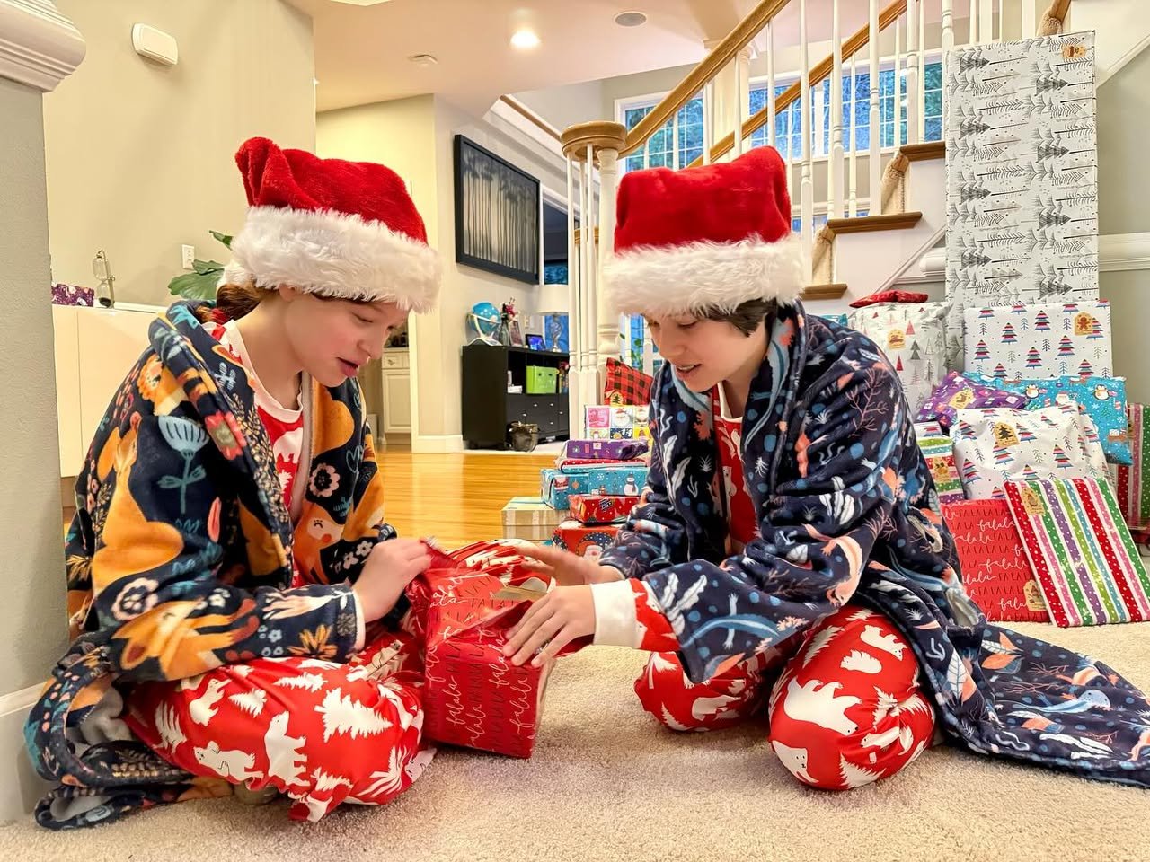 Two people wearing Santa hats and festive pajamas opening a gift in a home. Gift wrapping paper is stacked nearby.