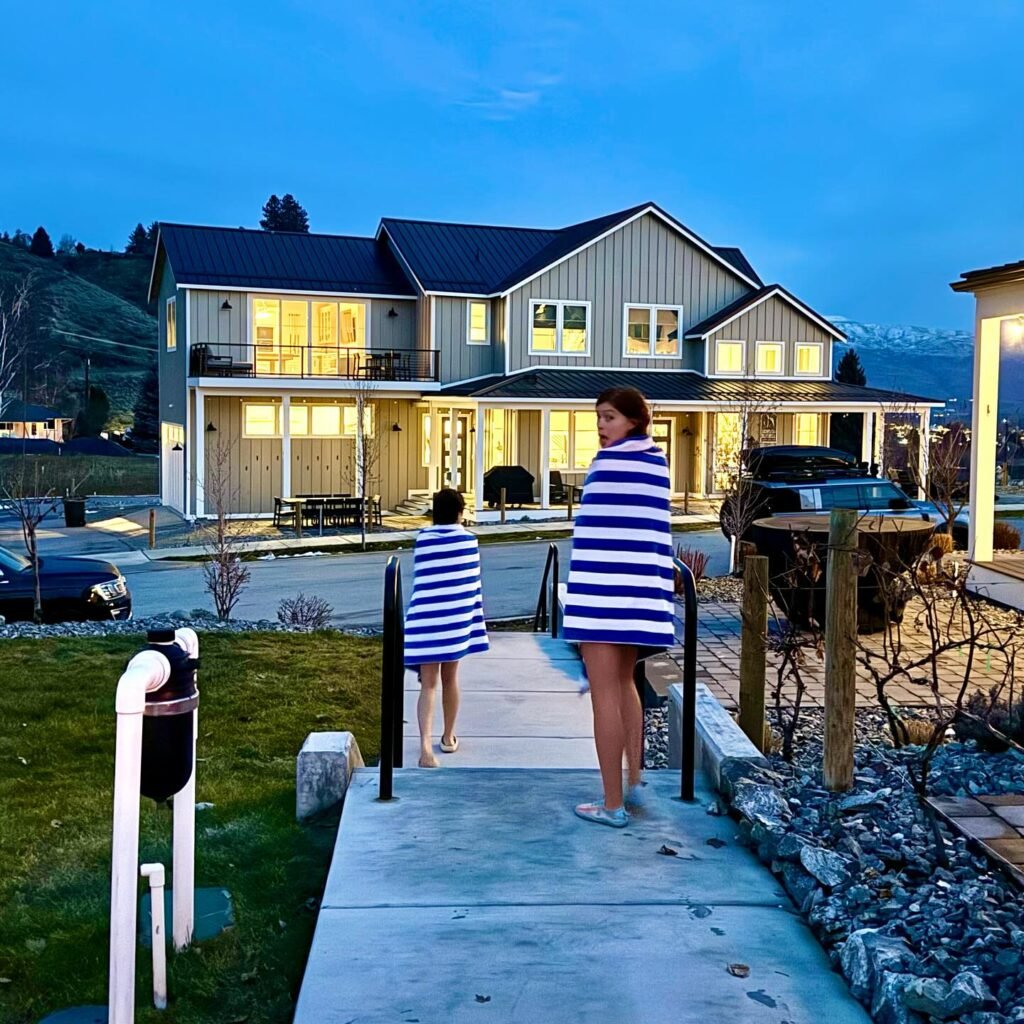 Two people wrapped in striped blankets walk toward a warmly lit two-story home at twilight.