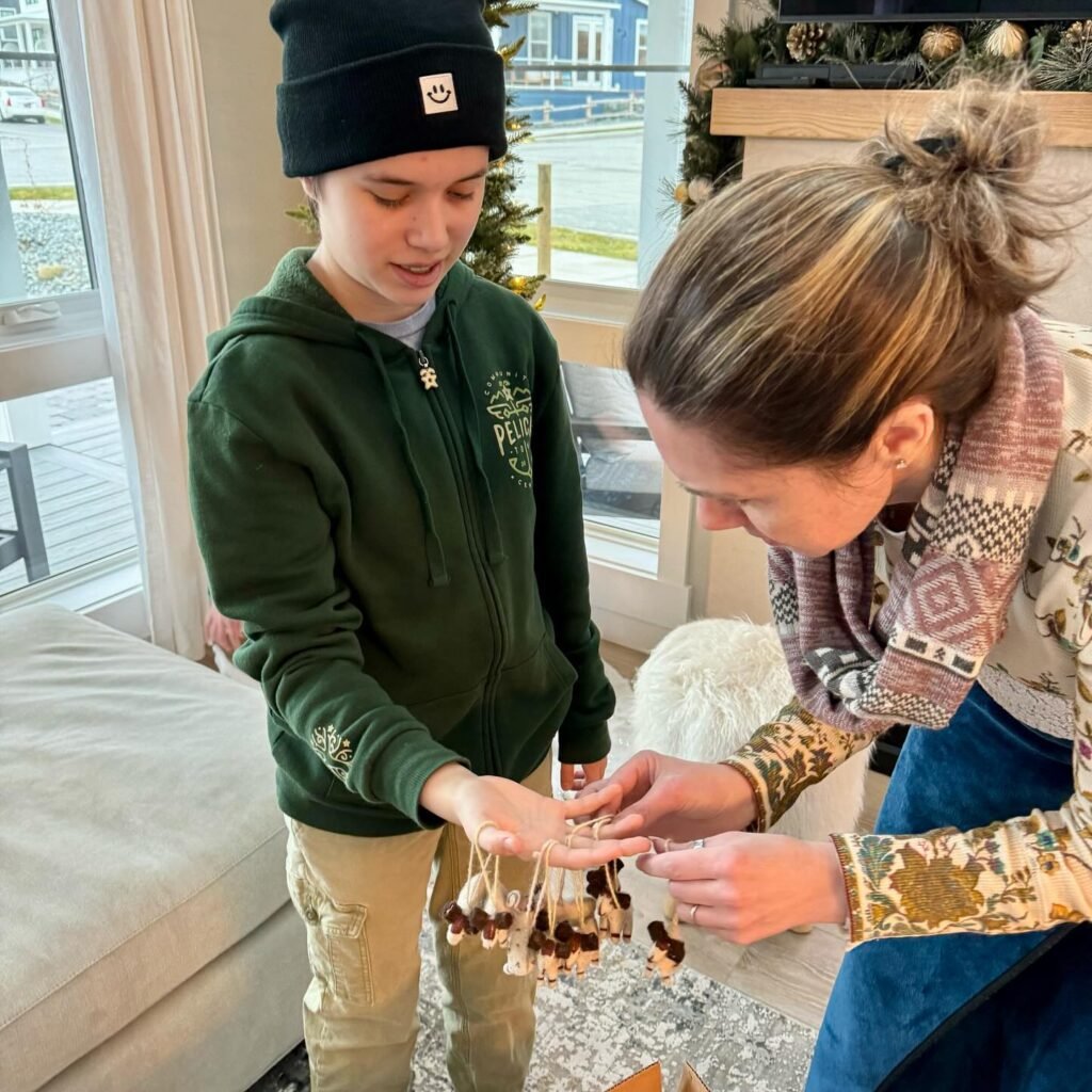 A young person in a green hoodie and beanie and another individual with a patterned sweater are close-up working on and handling small wooden or bark holiday ornaments.