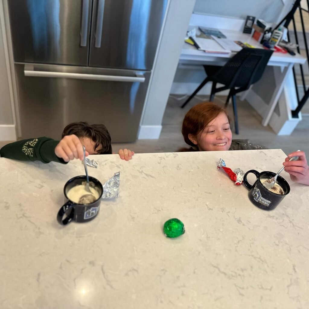 A child and a smiling person share a breakfast moment at a modern kitchen island. Hot drinks and colorful candy are on the countertop.