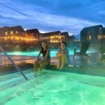 A glowing outdoor pool at dusk with two people relaxing on the edge, surrounded by illuminated resort buildings.