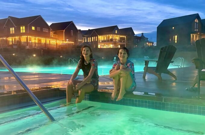 A glowing outdoor pool at dusk with two people relaxing on the edge, surrounded by illuminated resort buildings.