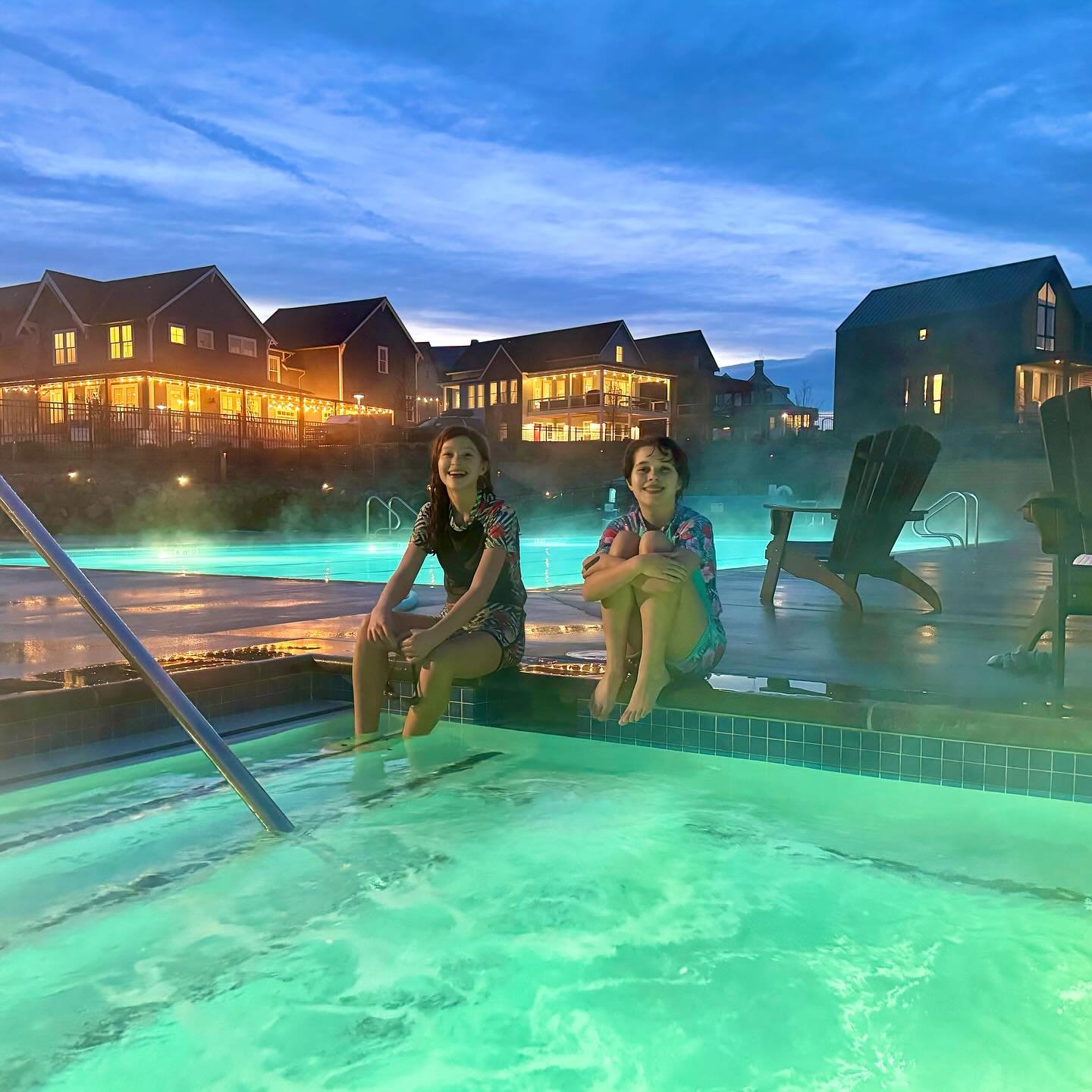 Happy New Year from Chelan A glowing outdoor pool at dusk with two people relaxing on the edge, surrounded by illuminated resort buildings.