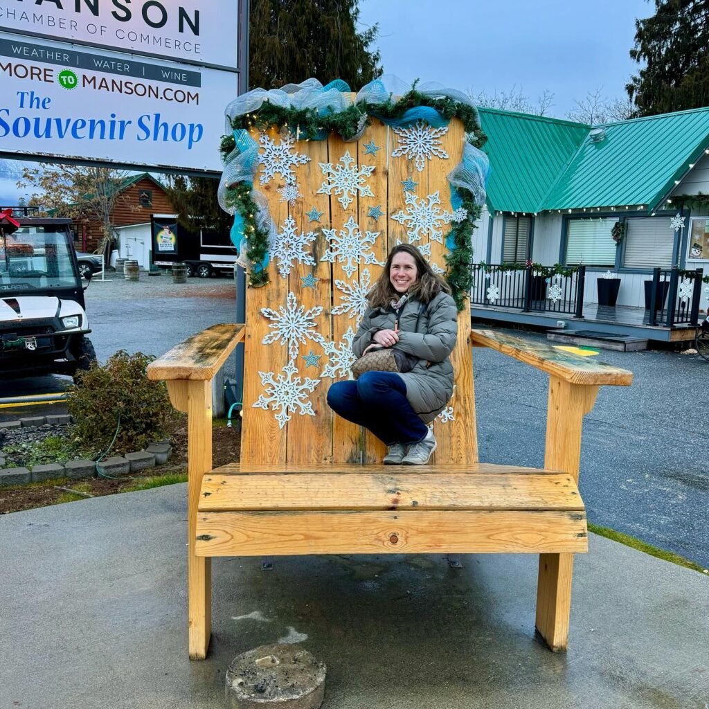 A person sits on a giant wooden chair decorated with white snowflakes and blue garlands. The chair is located outside a souvenir shop.