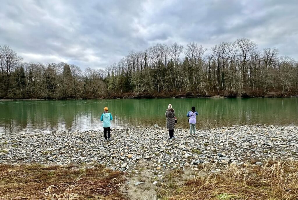 Visitors enjoy a stroll on a pebbly lake shore beneath overcast skies in the Pacific Northwest. The scene captures a quiet moment by the water's edge with leafless trees in the background.