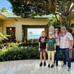 A group of four people smiling, wearing leis, posing in front of a building with a tropical ocean view.