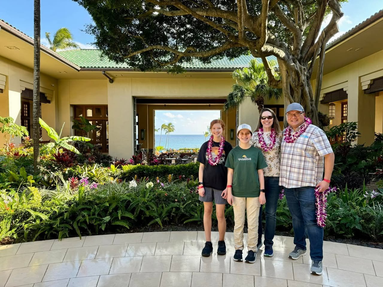 Grand Hyatt Kauai Resort & Spa A group of four people smiling, wearing leis, posing in front of a building with a tropical ocean view.