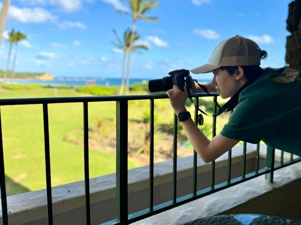 A person taking photos of a tropical landscape with palm trees and ocean from a balcony overlooking greenery.