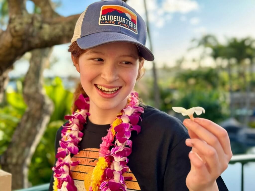 A smiling traveler wearing a Flamingo Park hat and a colorful flower lei holds a small white flower in a tropical setting.