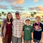 A family poses together at a tropical resort with palm trees and a pool in the background. The scene captures a beautiful sunset sky.