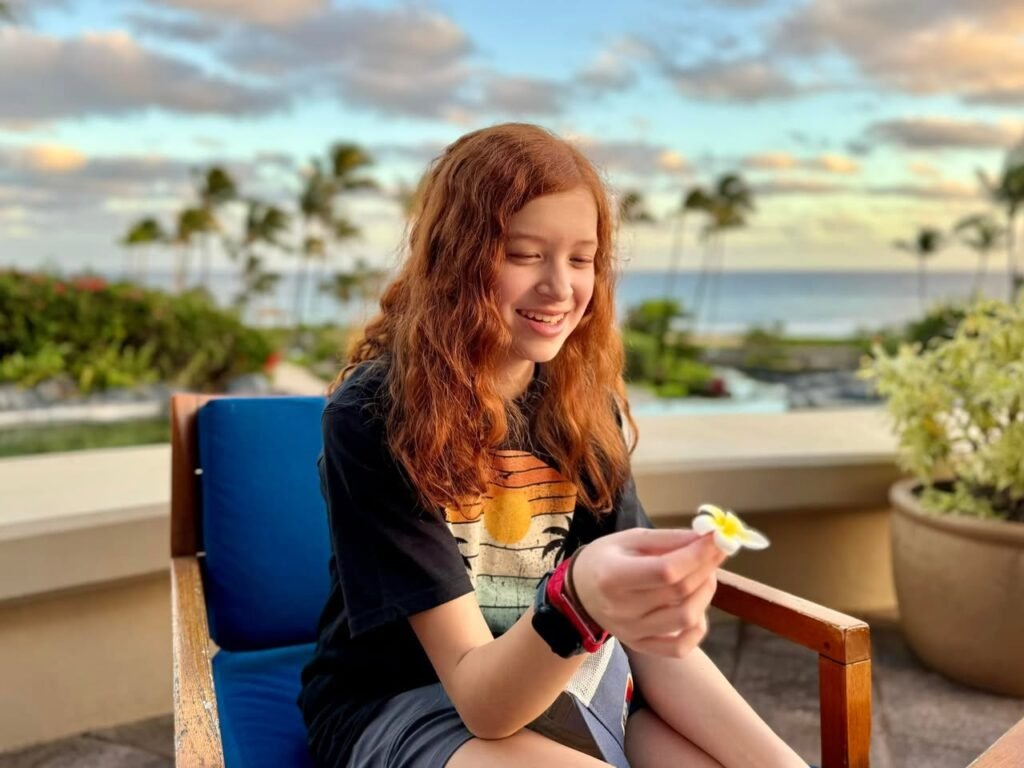 A person with red hair sits on a blue chair holding food against a tropical ocean backdrop. The scene captures a serene evening with palm trees and a soft sunset sky.