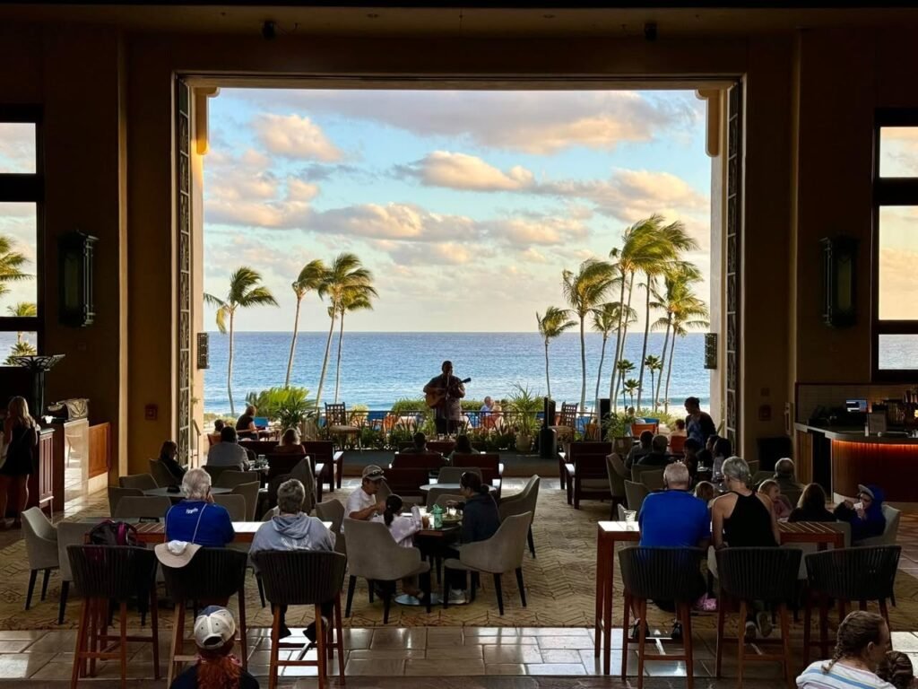 A tropical restaurant interior with guests dining while enjoying views of the ocean and swaying palm trees through an open door. Natural light from a bright sky illuminates the warmly decorated space.