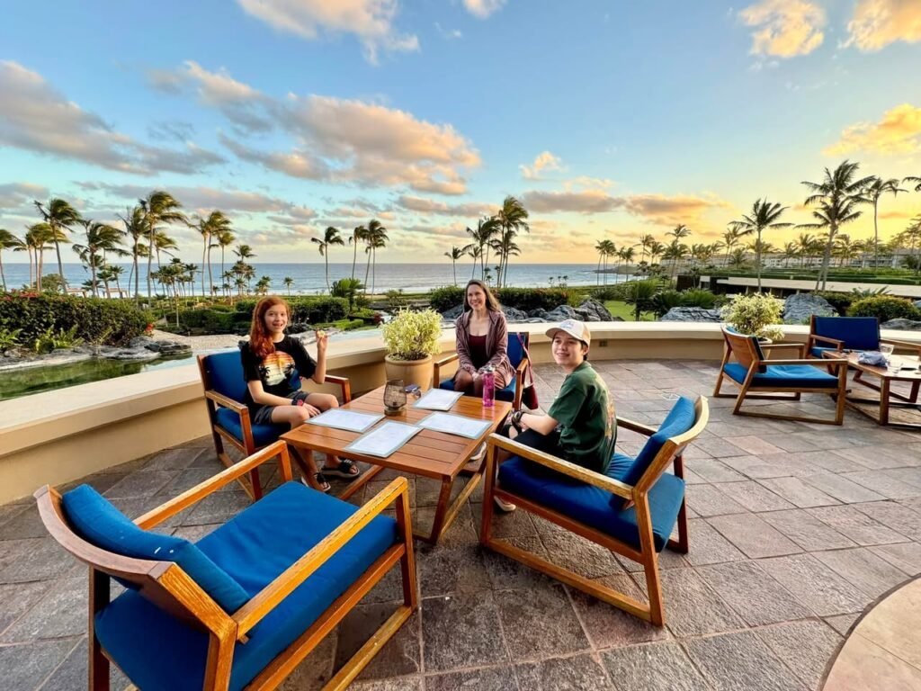 People enjoying a beachfront outdoor dining experience with palm trees and ocean views. Blue cushioned chairs on a stone patio.