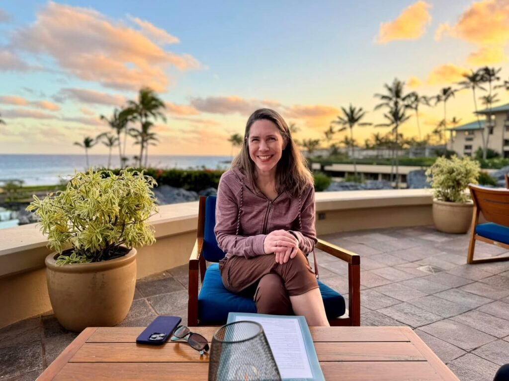 Person relaxes on a terrace overlooking the ocean and palm trees during golden hour. Reservations are being made at a nearby wooden table.