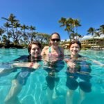 Family enjoying a tropical pool with palm trees and a clear blue sky background.