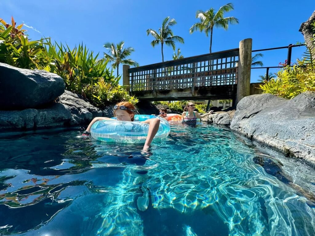 Floating on an inflator in a clear blue pool surrounded by lush foliage and a wooden rail.