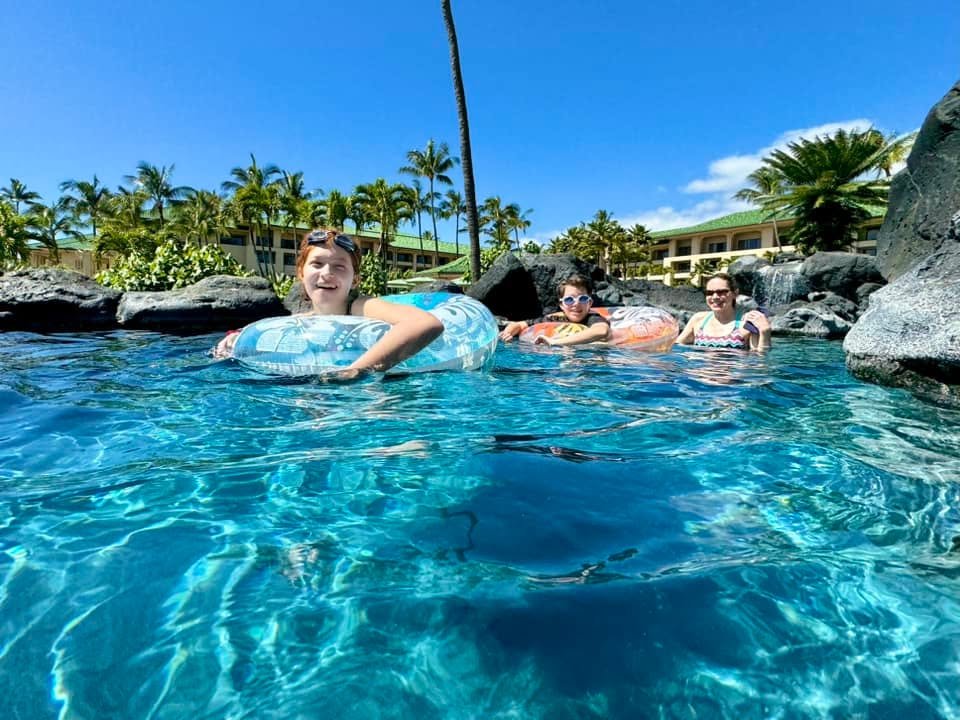 A person floats on a pool noodle in a clear, turquoise pool surrounded by tropical palms. Another individual relaxes nearby under a sunny sky.