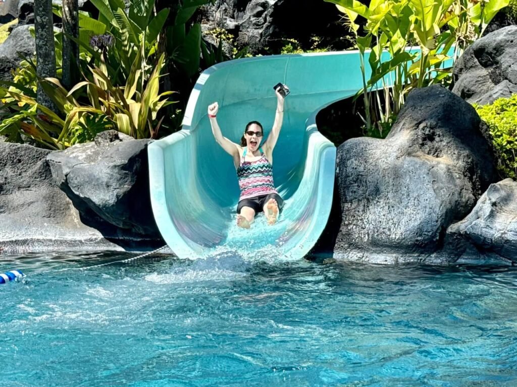 Excited person sliding into a pool on a green water slide surrounded by rocks and tropical plants.