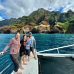 A family enjoying a catamaran vacation with a dramatic mountainous coastline in the background.