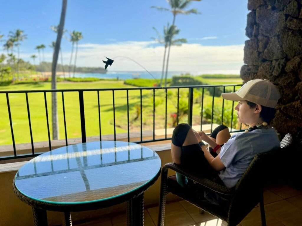 A person sits at a balcony table with a glass top, enjoying a tropical view featuring palm trees and the ocean. A stone wall is in the foreground of the area.