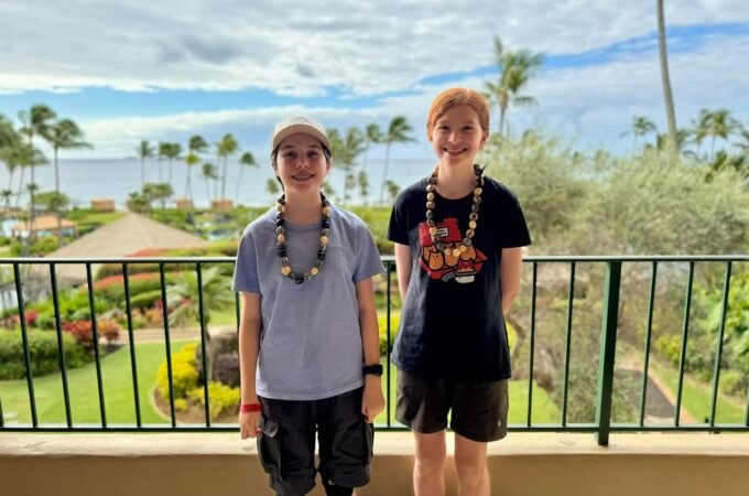 Two people standing on a balcony overlooking a tropical landscape with palm trees and ocean. The scene includes resort grounds and a clear blue sky.