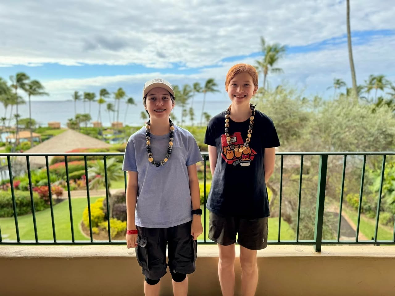 Kukui Nut Leis Two people standing on a balcony overlooking a tropical landscape with palm trees and ocean. The scene includes resort grounds and a clear blue sky.
