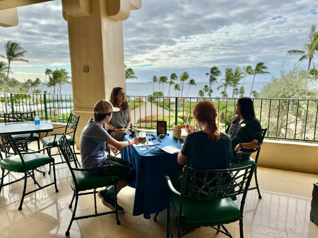 Diners sit at an outdoor cafe table surrounded by palm trees and a view of the ocean.