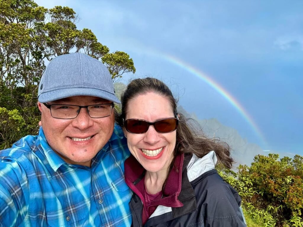A smiling couple takes a selfie in front of a double rainbow. They are outdoors with trees in the background.