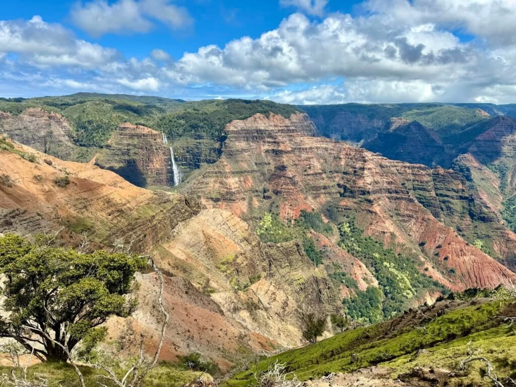 A dramatic view of Nahuku Fork from Kalalau Valley on the Na Pali Coast in Kauai, featuring waterfalls cascading from steep, layered cliffs under a bright blue sky.