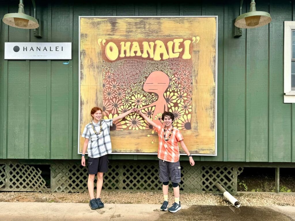 Travelers posing in front of a colorful Ohana Lei sign at a green building in Hanalei. Close-up views of the sign and lanterns on the building's facade.