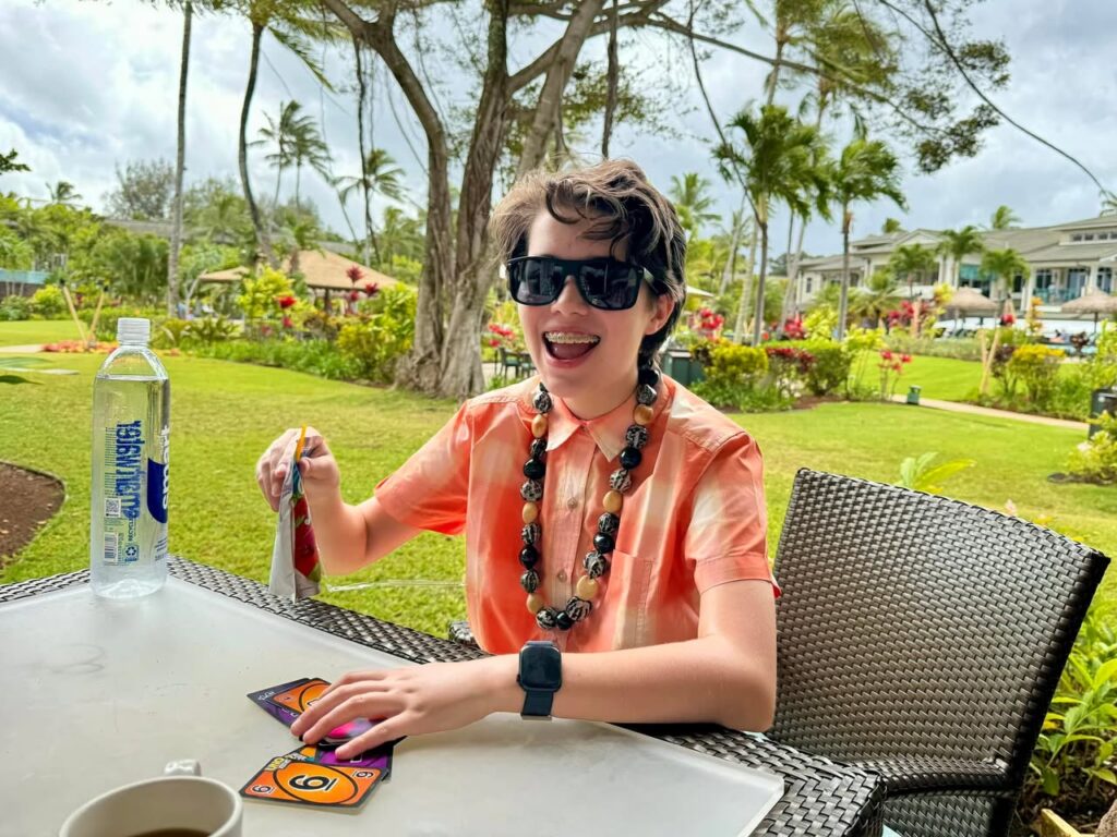 A smiling traveler playing cards at a sunny outdoor table with palm trees in the background. Tropical scenery surrounds a happy break.