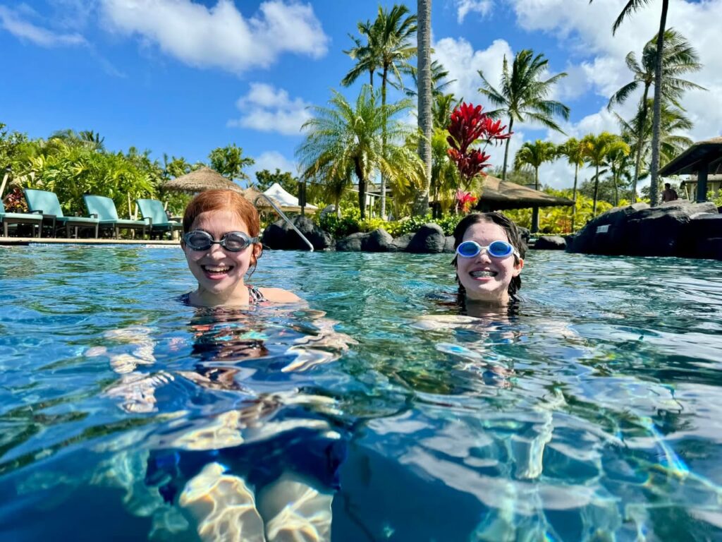 Two people relaxing in a resort pool surrounded by panoramic views of palm trees and a bright blue sky.