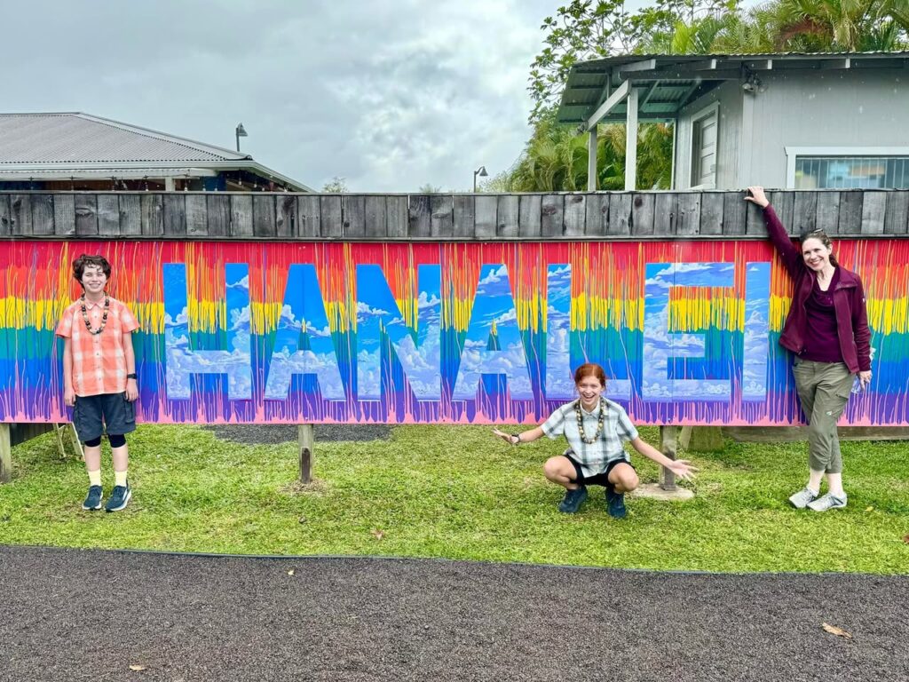 A group posing in front of a vibrant mural spelling out Hanalei on a wooden fence.