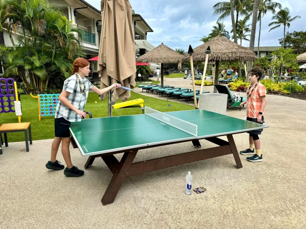 Two people playing table tennis on a resort patio with thatched umbrellas and palm trees.