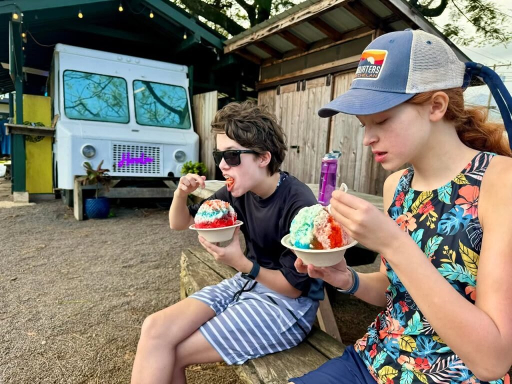 Two people enjoying ice cream outside while sitting on a bench. In the background, there is a van and a small building.