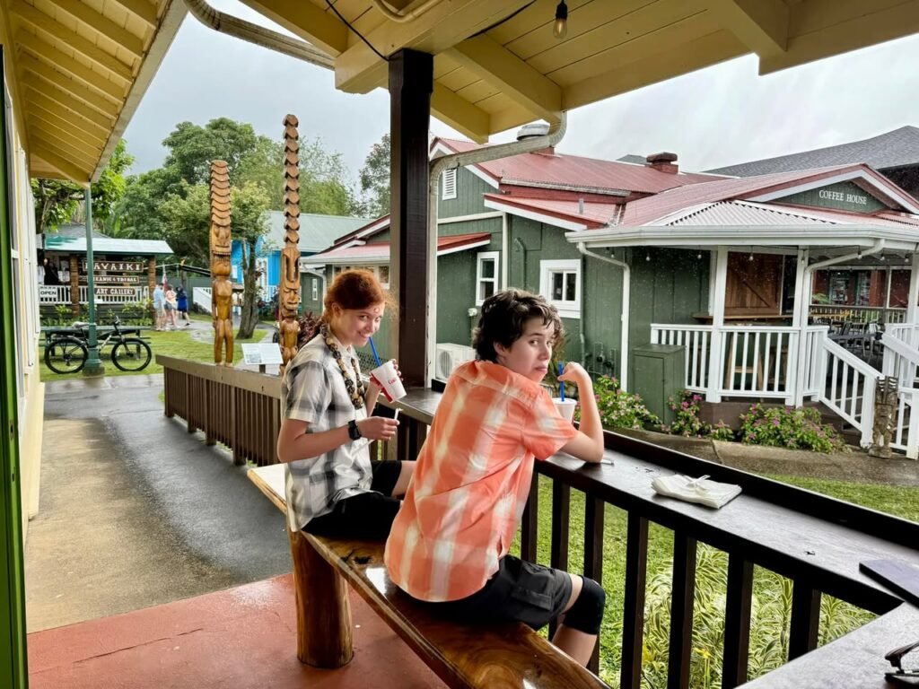A view of a campground featuring a wooden porch and surrounding A-frame cabins with green walls. People sit on the bench watching the scenery.