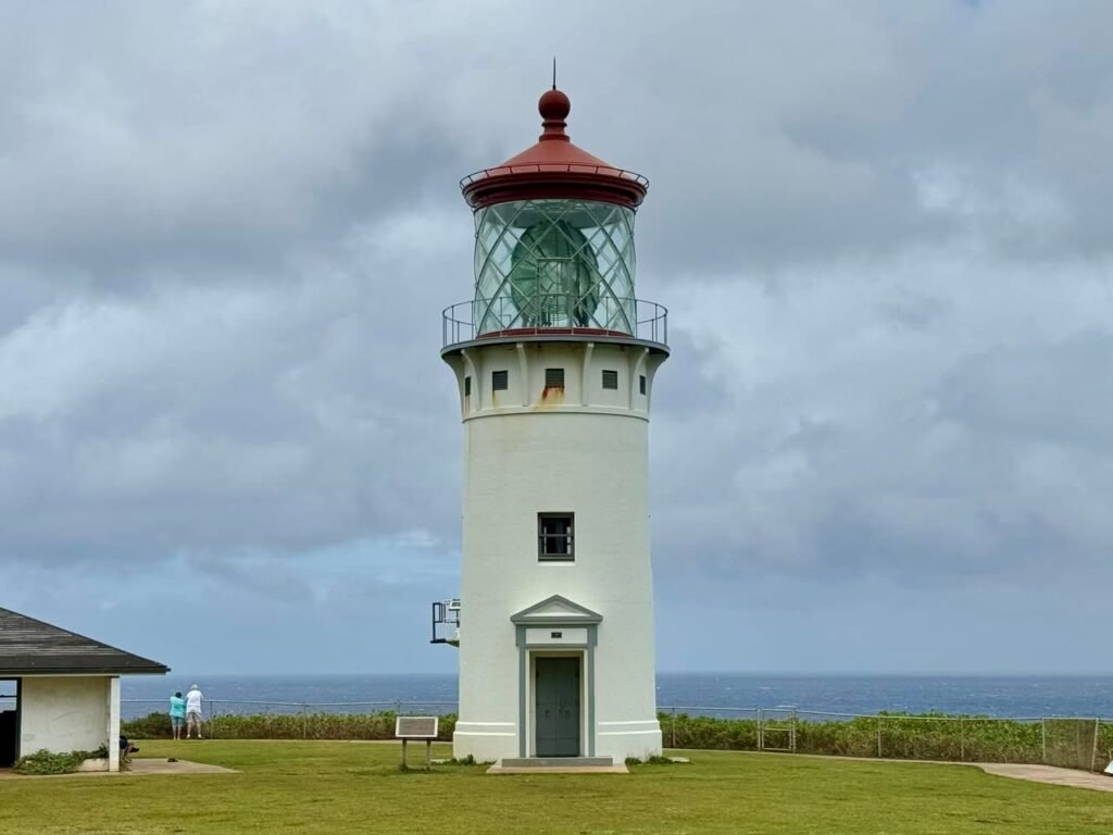 A white lighthouse with a red roof standing near the sea under a cloudy sky. Close up details of the lighthouse lens and a patio area.