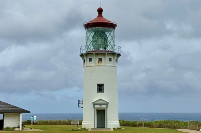 A white lighthouse with a red roof standing near the sea under a cloudy sky. Close up details of the lighthouse lens and a patio area.