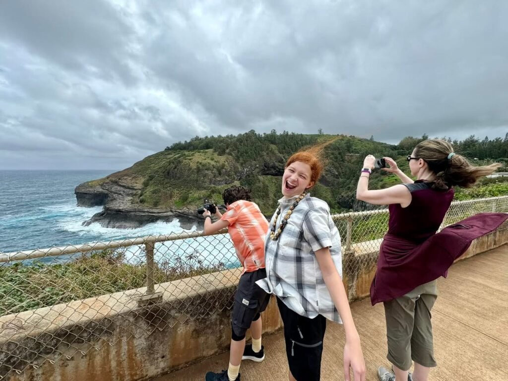 Travelers pose on a windy cliffside viewpoint under a dramatic, overcast sky near a rugged coastline.