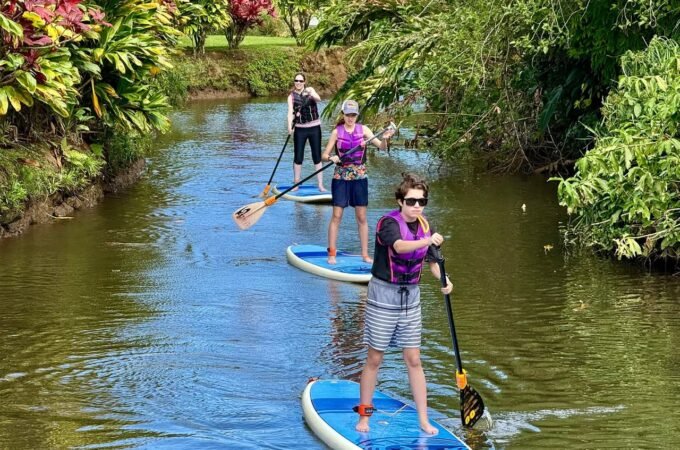 Three people paddleboard down a lush canal in vibrant life jackets, surrounded by tropical foliage. This serene scene captures a family-friendly outdoor adventure.
